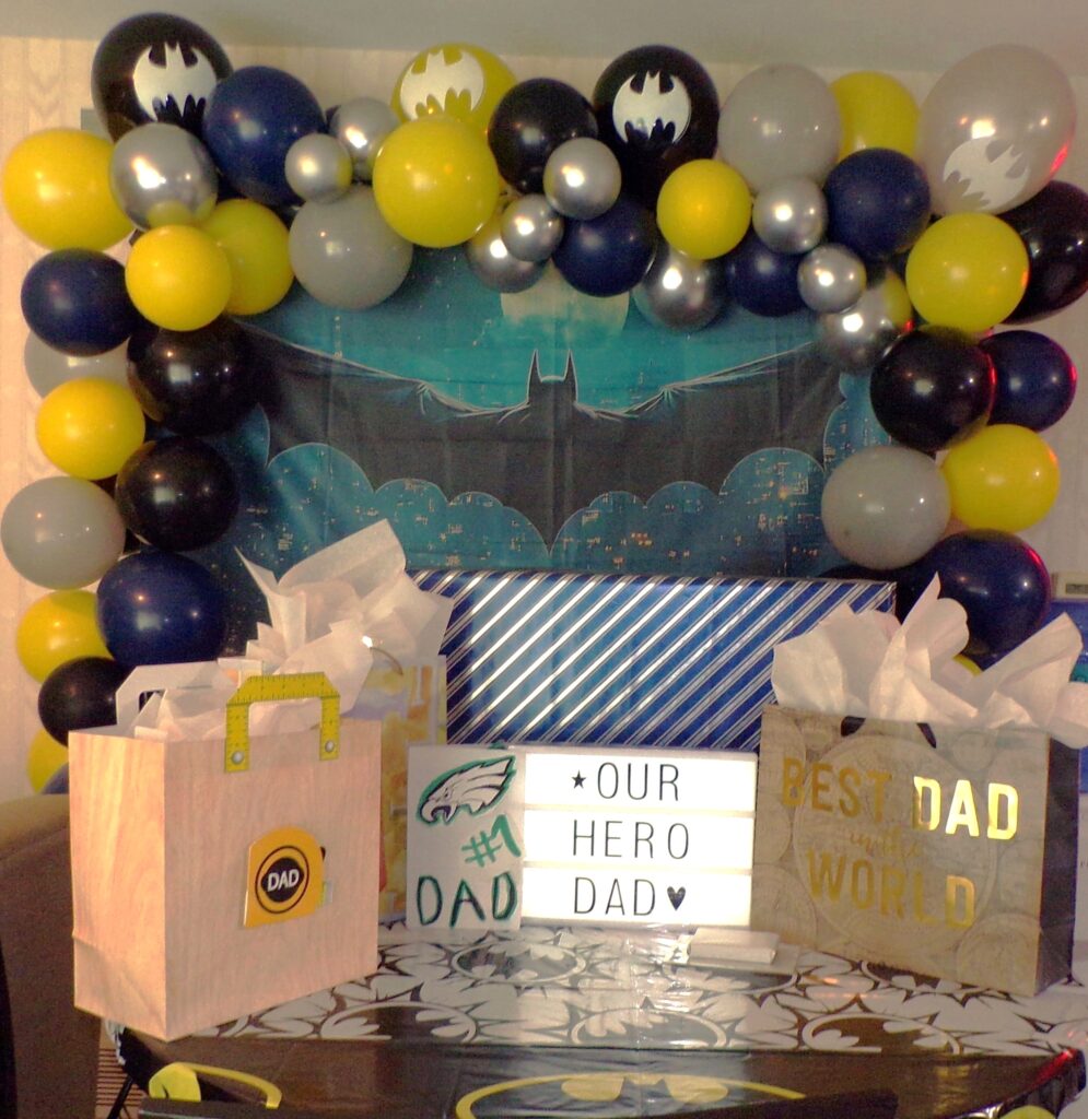 Bat  Backdrop with black, blue, grey, yellow and silver balloon arch with batman balloon accents and Father's Day gift table setup